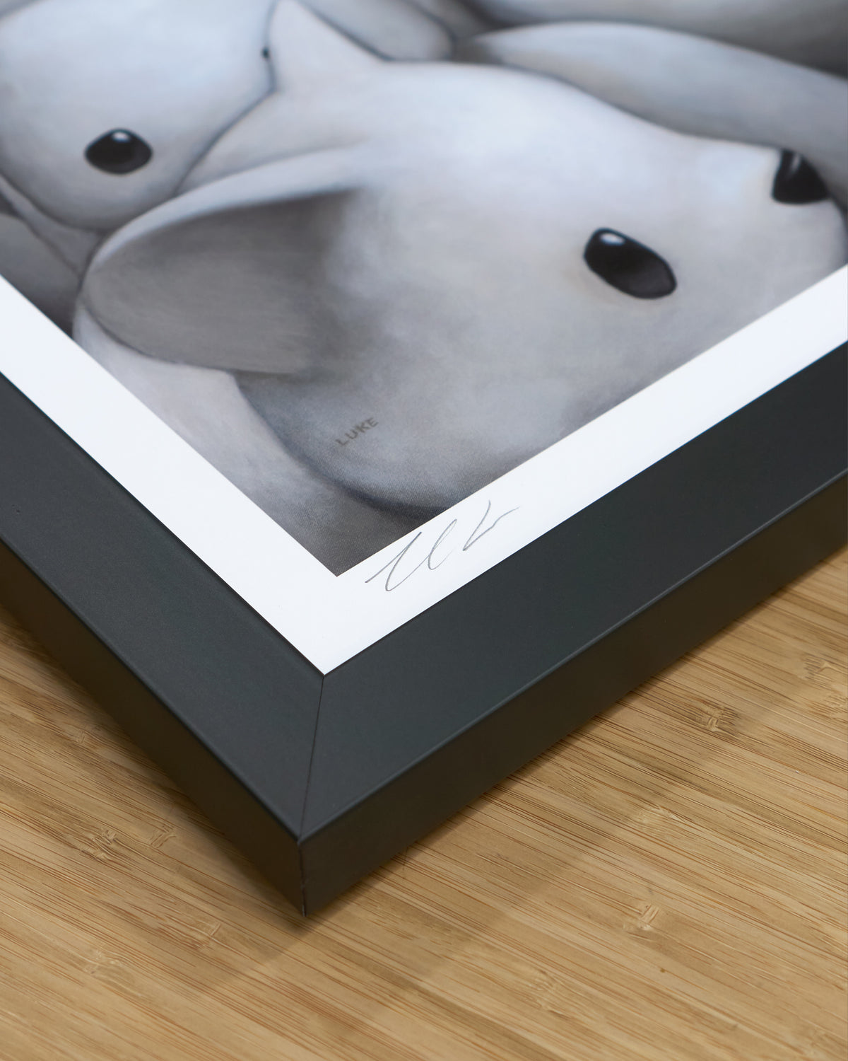 A framed print showing a close-up of frame corner and Luke Chueh's signature, photographed at an angle on a light wood floor. The frame is dark charcoal gray. 
