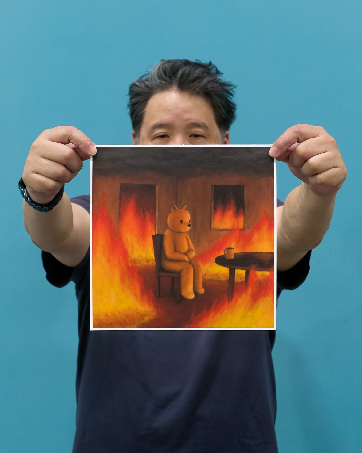 An expressive painting of a small yellow bear-like figure sitting on a chair in a burning room, with flames filling the walls and floor and a mug on a table beside it.